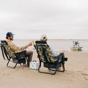Two friends relaxing on the beach in PARKIT × Free Fly Eclipse Recliners in Vintage Camo, with a Turtlebox speaker playing between them and a skiff anchored in the background. These premium folding outdoor chairs feature a built-in cooler, backpack straps, and recline function perfect for beach days, boat trips, or backyard hangs for dads, moms, and outdoor enthusiasts alike.