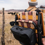 Person with a patterned chair and black bag in a field with cows in the background